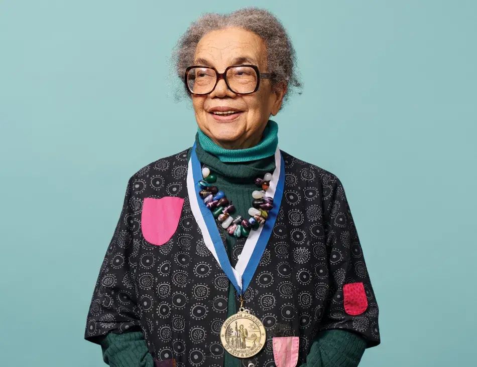 A headshot of Marian Wright Edelman, an elderly woman with brown skin and curly gray hair parted in the middle, as she faces the camera and smiles. Edelman wears two blue turtlenecks, a black and gray sweater, and a colorful beaded necklace. Photo by Noah Willman.